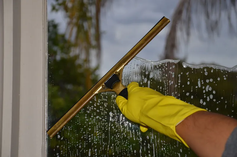 Hand im gelben Handschuh zieht mit einem Abzieher Schaum und Wasser von einer Fensterscheibe